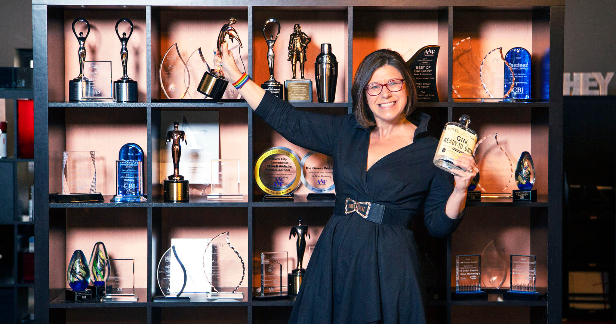 wide shot of Jen Neumann standing in front of a shelf with rewards on it and holding a bottle of gin