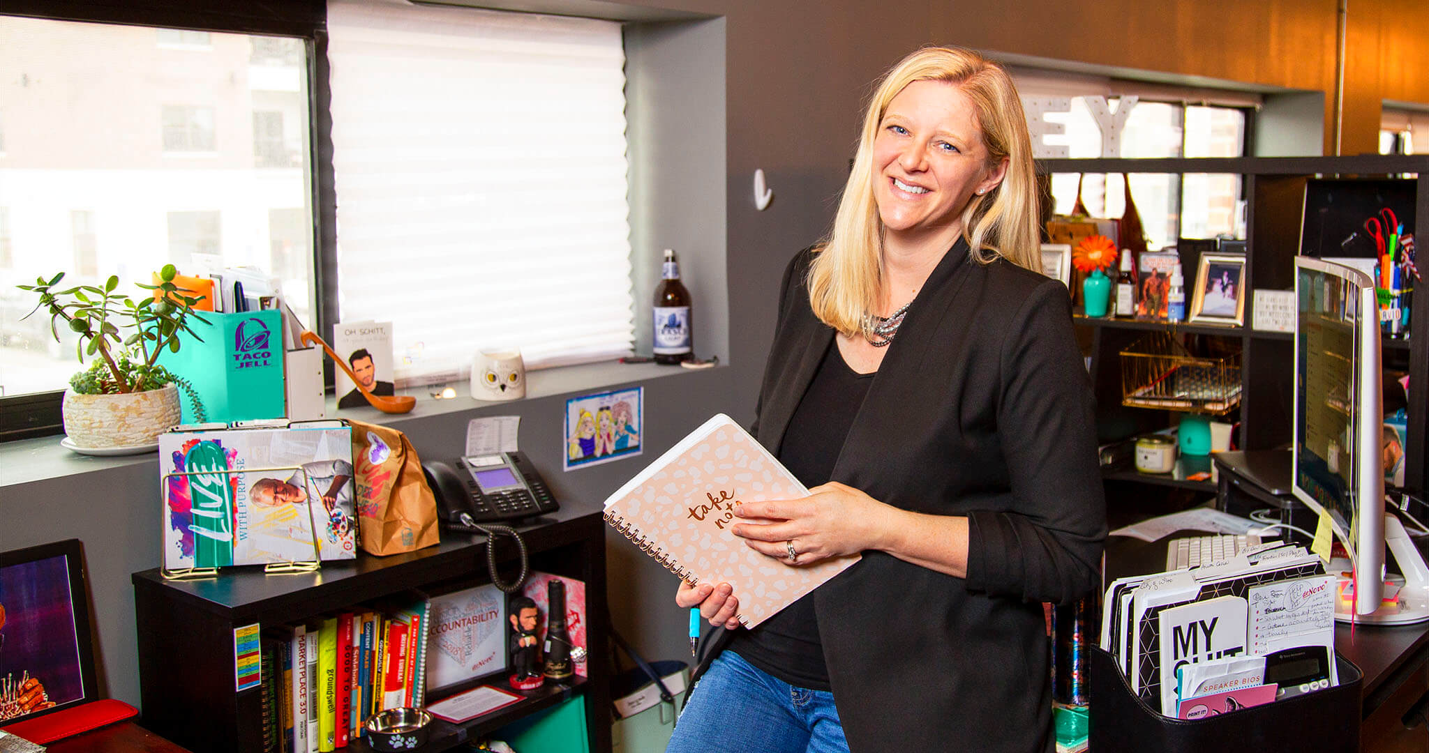 wide shot of Jill Mast leaning on her desk holding a notebook and smiling
