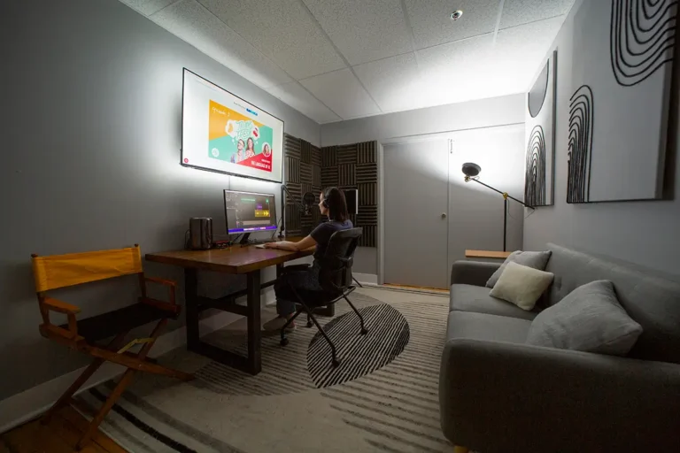 Modern office studio space with person working at desk, featuring monitors, acoustic panels, couch, and professional lighting setup