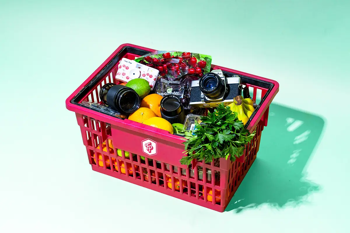 Bright pink shopping basket overflowing with cameras, fresh produce, and herbs against a turquoise background, representing a marketing agency's blend of creative tools and fresh ideas