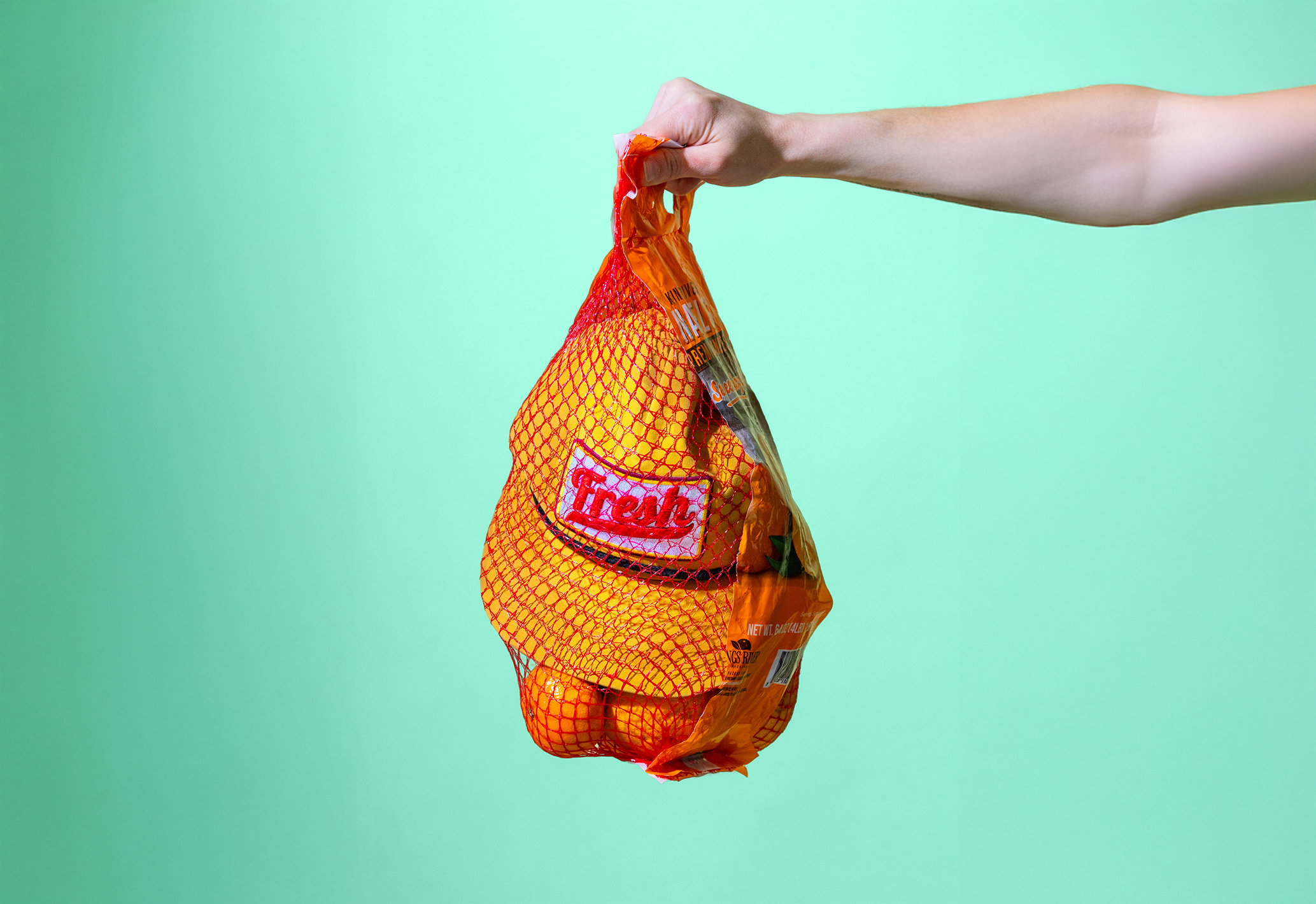 Hand holding a mesh bag of oranges with a product label against a turquoise background
