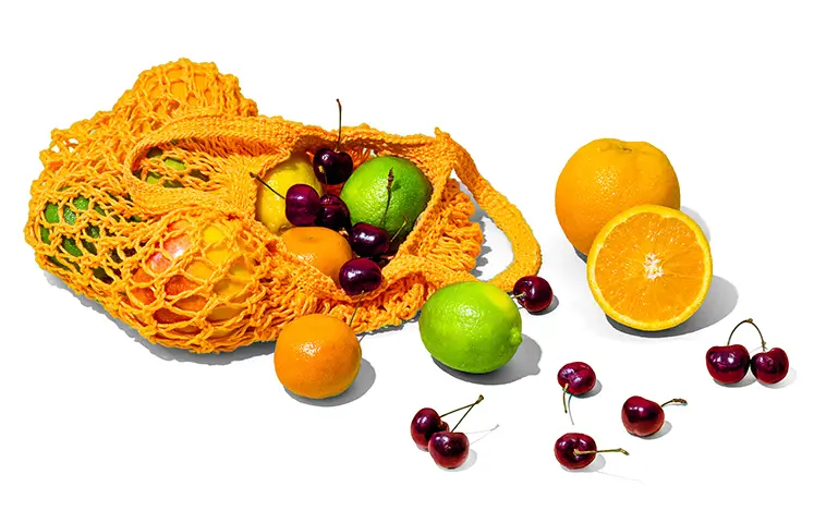 Colorful fresh fruits including oranges, limes, and cherries spilling from a yellow mesh produce bag, representing fresh, natural ingredients