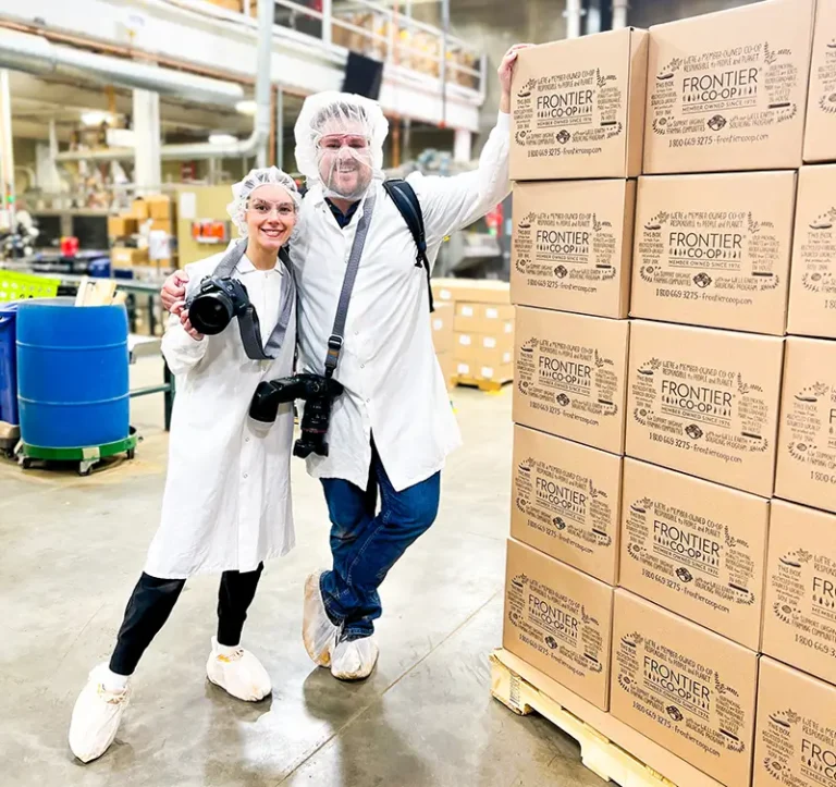 Two employees in white lab coats and hairnets celebrate in a production facility, holding cameras in front of stacked cardboard boxes labeled Frontier
