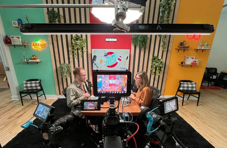 Two people recording a podcast or video interview in a colorful studio with professional cameras, monitors, and microphones on a wooden desk