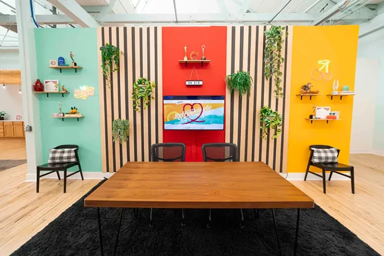 Podcast recording space styled with colorful accent walls in mint, red, and yellow, featuring wooden desk, striped wood paneling, potted plants, shelving, and a mounted TV screen