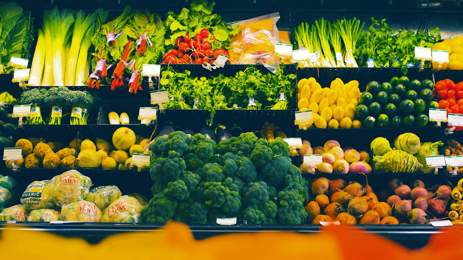 Vibrant farmers market display with fresh produce including broccoli, peppers, lettuce, leeks, citrus fruits, and root vegetables arranged on tiered shelves