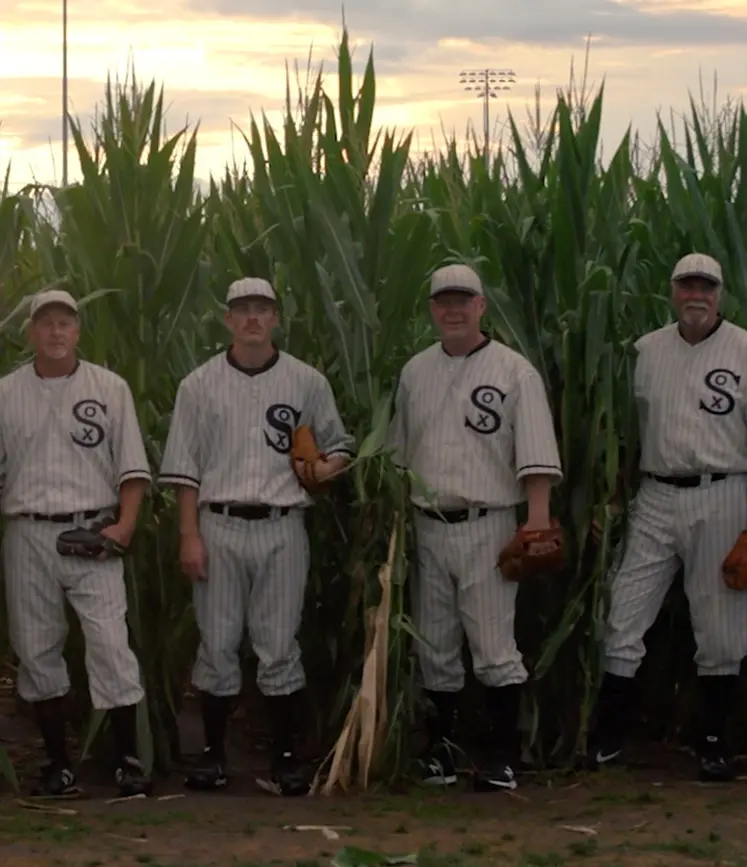 Four baseball players in vintage Chicago White Sox uniforms stand in a cornfield at dusk, holding baseball gloves